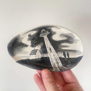 以下が含まれることがあります： UFOが農家に光線を当てている白黒イラストが描かれた楕円形のセラミック皿。皿は濃い灰色の縁と光沢のある仕上げが施されています。このアートワークは、嵐の空の下の田園風景を描いています。