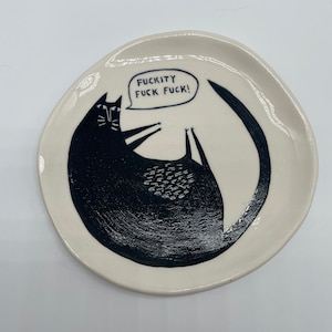 Może przedstawiać: Mały, kremowy ceramiczny talerz z czarną ilustracją kota. Kot leży na plecach z ogonem wok&oacute;ł niego. Dymek nad kotem m&oacute;wi "FUCKITY FUCK FUCK!" czarnymi literami.