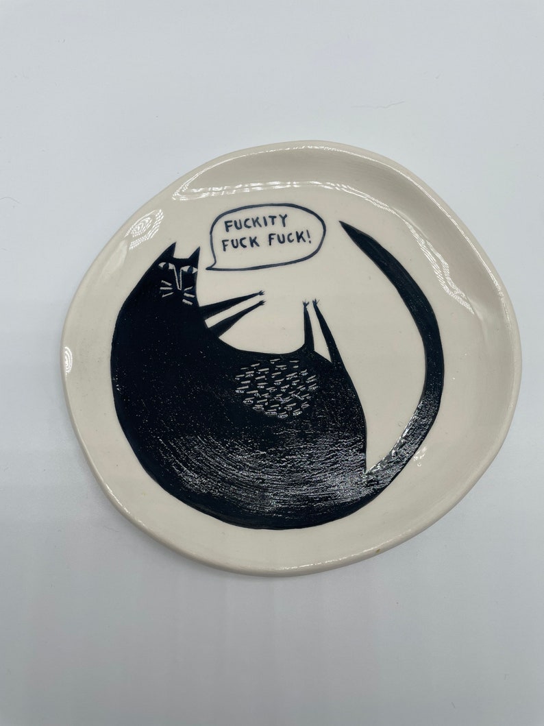 Może przedstawiać: Mały, kremowy ceramiczny talerz z czarną ilustracją kota. Kot leży na plecach z ogonem wok&oacute;ł niego. Dymek nad kotem m&oacute;wi "FUCKITY FUCK FUCK!" czarnymi literami.