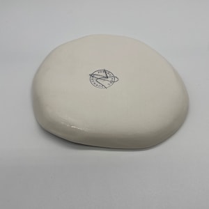 Może przedstawiać: Biały, nieregularny ceramiczny talerz. Sp&oacute;d posiada okrągłą pieczęć z napisem "JOE RUTHERS HANDMADE". Talerz ma gładkie, błyszczące wykończenie i minimalistyczny design.