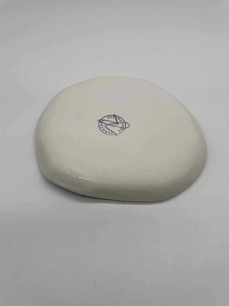 Może przedstawiać: Biały, nieregularny ceramiczny talerz. Sp&oacute;d posiada okrągłą pieczęć z napisem "JOE RUTHERS HANDMADE". Talerz ma gładkie, błyszczące wykończenie i minimalistyczny design.