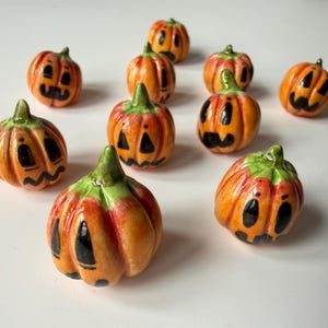 Könnte beinhalten: Eine Sammlung von zehn kleinen Kürbissen aus Keramik, jeweils mit glänzend orangefarbener Oberfläche, grünen Stielen und handbemalten schwarzen Jack-o'-Lantern-Gesichtern. Die Kürbisse variieren leicht in Größe und Ausdruck und zeigen verschiedene Schnitzmuster.