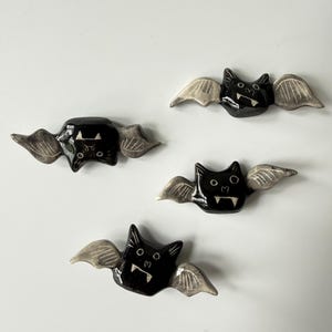 May include: Four small, handcrafted ceramic bat figurines arranged in a cluster. Each bat features a glossy black body with white painted details, including pointed ears, circular eyes, and two sharp fangs. The wings are light gray with etched lines.