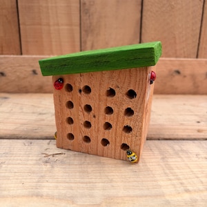 May include: A wooden insect hotel featuring a slanted green roof and a front panel with sixteen drilled holes. The natural wood structure is decorated with small, colorful ladybug and bee figurines attached to the corners of the front face.