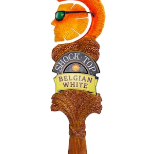 May include: A Shock Top Belgian White beer tap handle featuring a stylized orange slice character with a mohawk and green sunglasses. The base is shaped like a bundle of wheat stalks with a dark brown plaque displaying the brand name and text.