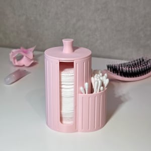 Puede incluir: Un organizador cilíndrico de plástico rosa claro con una ranura vertical llena de una pila de discos de algodón redondos blancos y un compartimento abierto adyacente que contiene bastoncillos de algodón blancos, con un pequeño pomo central.