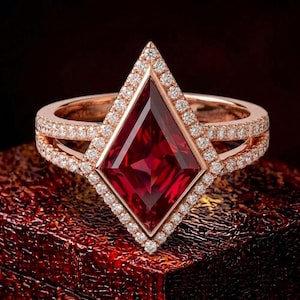 May include: A rose gold ring featuring a large, kite-shaped red gemstone at the center. The kite-cut stone is surrounded by a halo of small, round white diamonds. The split shank band is also encrusted with small, round white diamonds throughout.