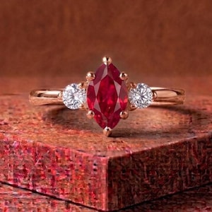 May include: A rose gold ring featuring a central marquise-cut red gemstone flanked by two round-cut clear stones. The band is smooth and polished, holding the three-stone setting securely in place with metal prongs on a textured surface.