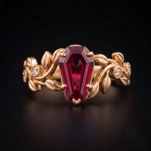 May include: A gold ring featuring a central coffin-cut deep red gemstone. The band is designed with sculpted gold leaves and vines, accented by small round white stones embedded within the metal foliage on both sides of the center stone.