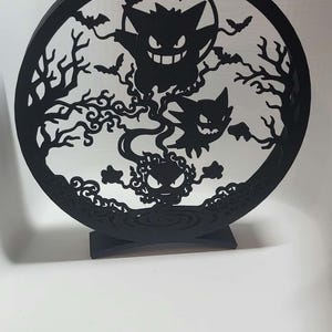 May include: A circular black laser-cut display piece featuring three ghost-like creatures, bare tree branches, and small bats. The design is enclosed within a round frame and is supported by a curved base, creating a detailed silhouette scene.