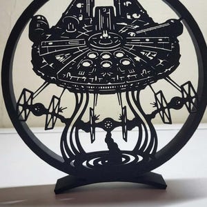 May include: A black circular decorative stand featuring a detailed laser-cut silhouette of a starship resembling the Millennium Falcon. The design includes smaller starfighter silhouettes suspended below the main ship, all enclosed within a ring.