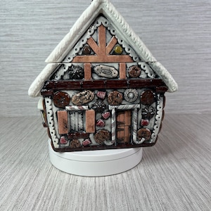 May include: A ceramic gingerbread house figurine featuring a white snow-dusted roof, brown walls, and decorative elements resembling cookies, candies, and icing. The structure includes a central door, a window with shutters, and a chimney on top.