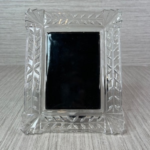 May include: A rectangular clear glass picture frame featuring a decorative cut pattern of repeating leaf motifs along the border. The frame has a solid black rectangular center insert and sharp, faceted corners with a polished finish.