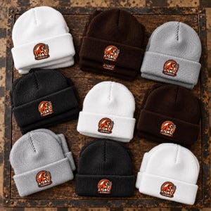 May include: A collection of nine knit beanies in white, brown, gray, and black. Each beanie features a cuffed brim and a circular patch displaying an orange and white bulldog graphic with the text "OLD DOG SUPPLY CO" printed in a circular border.