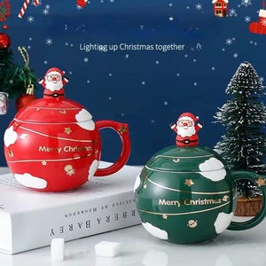 Può includere: Due tazze rotonde in ceramica con coperchio, una rossa e una verde, ciascuna con una statuetta di Babbo Natale sulla parte superiore, motivi a nuvola bianchi, dettagli a stella dorati e la scritta Merry Christmas in lettere dorate.