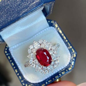 Pode incluir: Um anel com uma grande pedra central de rubi vermelho de corte oval rodeada por um halo de múltiplos diamantes brancos de corte pera e redondo. O anel está montado numa banda metálica e é apresentado dentro de uma caixa de joias azul escura.