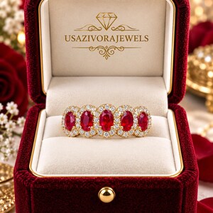 May include: A gold ring featuring five oval-cut red rubies arranged in a row, each surrounded by a halo of small white diamonds. The ring is displayed inside a deep red velvet jewelry box with the text USAZIVORAJEWELS printed in gold on the lid.