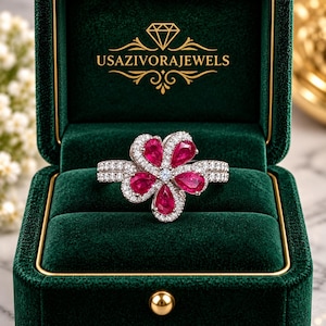 Pode incluir: Um anel prateado com design de flor, apresentando cinco rubis vermelhos em corte de pera rodeados por pequenas pedras brancas. O anel é exibido dentro de uma caixa de veludo verde-escura com o texto USAZIVORAJEWELS impresso em dourado na tampa.