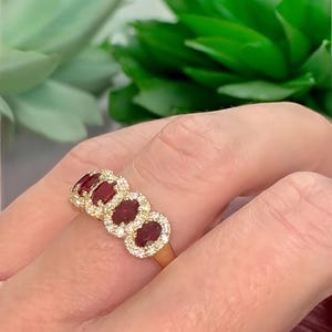 Peut inclure: Une bague en or orn&eacute;e de quatre rubis rouge fonc&eacute; taille ovale dispos&eacute;s en rang&eacute;e horizontale. Chaque rubis est entour&eacute; d'un halo de petits diamants blancs ronds et transparents, mont&eacute;s sur un anneau en or poli entourant le doigt.