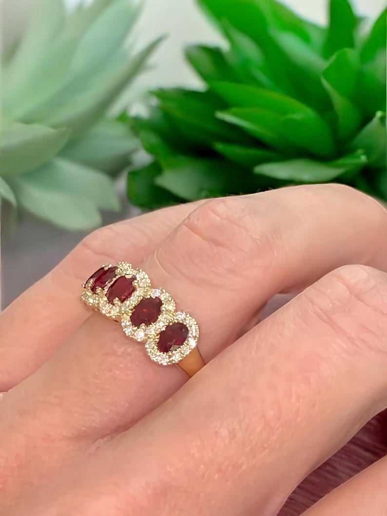 Peut inclure: Une bague en or orn&eacute;e de quatre rubis rouge fonc&eacute; taille ovale dispos&eacute;s en rang&eacute;e horizontale. Chaque rubis est entour&eacute; d'un halo de petits diamants blancs ronds et transparents, mont&eacute;s sur un anneau en or poli entourant le doigt.