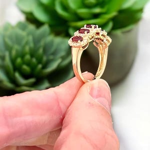 Peut inclure: Une bague en or orn&eacute;e de trois pierres pr&eacute;cieuses rouge fonc&eacute; de taille ovale, dispos&eacute;es en rang&eacute;e et entour&eacute;es chacune d'un halo de petites pierres blanches. L'anneau est en or poli, pr&eacute;sentant un design classique avec un sertissage.