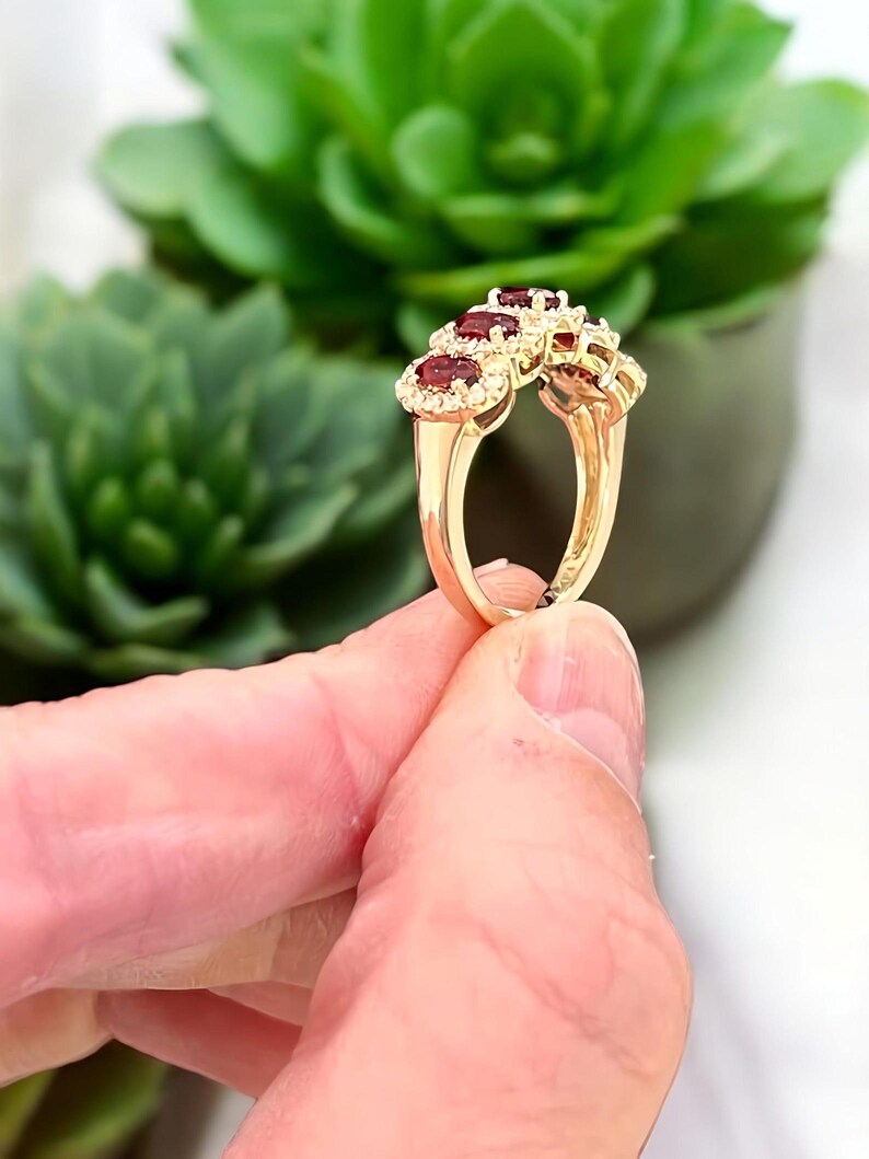 Peut inclure: Une bague en or orn&eacute;e de trois pierres pr&eacute;cieuses rouge fonc&eacute; de taille ovale, dispos&eacute;es en rang&eacute;e et entour&eacute;es chacune d'un halo de petites pierres blanches. L'anneau est en or poli, pr&eacute;sentant un design classique avec un sertissage.