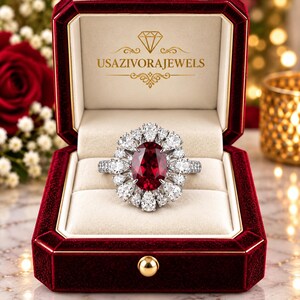 Puede incluir: Un anillo con una gran piedra preciosa de color rojo intenso en talla ovalada rodeada por un halo de piedras claras en forma de pera, presentado en un estuche de terciopelo rojo oscuro con el texto USAZIVORAJEWELS impreso en oro en la tapa.