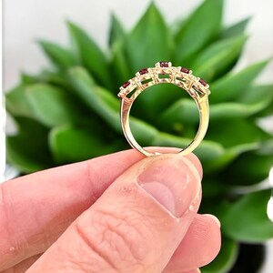 Peut inclure: Une bague en or orn&eacute;e d'une rang&eacute;e de cinq petites pierres pr&eacute;cieuses rondes d'un rouge profond, serties le long de l'anneau sup&eacute;rieur. L'anneau en m&eacute;tal est poli et lisse, pr&eacute;sentant un sertissage &agrave; griffes classique pour chaque pierre.