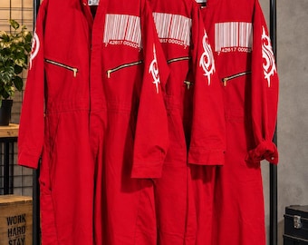 Pode incluir: Três macacões vermelho-brilhantes pendurados em cabides brancos. Cada macacão apresenta um gráfico de código de barras branco no peito com o número 42617 00002 abaixo, dois bolsos horizontais com zíper no peito e um design tribal branco nas mangas.