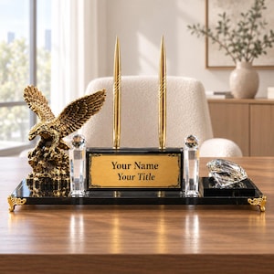 Pode incluir: Um conjunto de placa de identificação de mesa com base retangular preta e pés dourados, uma figura de águia dourada, duas canetas douradas, dois pilares de cristal transparente e um diamante de cristal facetado. Uma placa dourada exibe o texto Your Name Your Title.