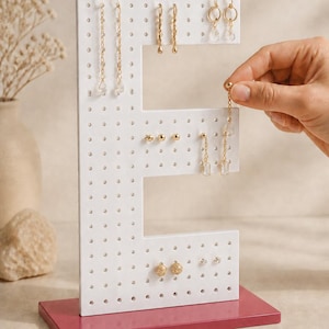 May include: A white, E-shaped earring display stand with a maroon rectangular base, featuring multiple rows of holes for organizing jewelry. The stand holds various gold-toned earrings, including drop, hoop, and stud styles, arranged neatly across the surface.