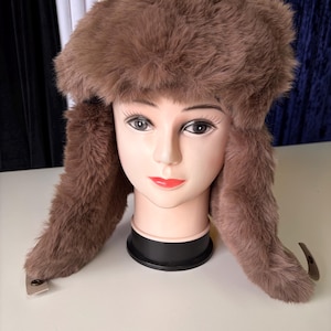 May include: A brown faux fur trapper hat with long ear flaps and small metallic snap fasteners at the ends. The hat features a thick, plush texture and is displayed on a mannequin head with neutral facial features and painted details.