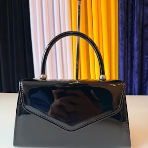 May include: A black patent leather handbag with a glossy finish, featuring a structured envelope-style flap, a single rounded top handle, and silver-toned metal hardware attachments connecting the handle to the main body of the bag.