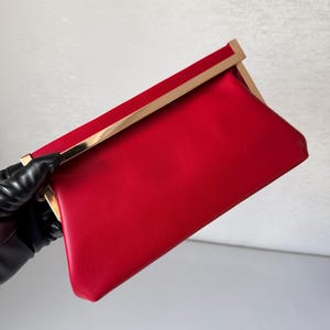 May include: A vibrant red rectangular clutch bag featuring a smooth, solid-coloured exterior and a polished gold-tone metal frame along the top edge. The accessory has a clean, structured design with a secure clasp closure and a sleek, minimalist finish.