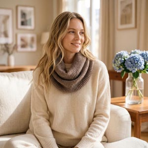 May include: A ribbed knit infinity scarf in a warm taupe brown colour, featuring a thick, textured wool-like material that wraps around the neck in multiple layers, paired with a long-sleeved, cream-coloured sweater with a simple crew neckline.