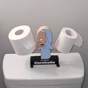 May include: A black toilet paper holder featuring a cutout of the character The Great Cornholio with a light blue shirt pulled over the head. The character has two extended arms that hold two rolls of white toilet paper. The base is black with white text.