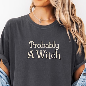 Può includere: Una t-shirt a maniche corte color grigio antracite con il testo "Probably A Witch" stampato in un carattere serif color crema sul petto. La maglietta ha uno scollo rotondo ed è indossata con una giacca di jeans azzurra sulle spalle.