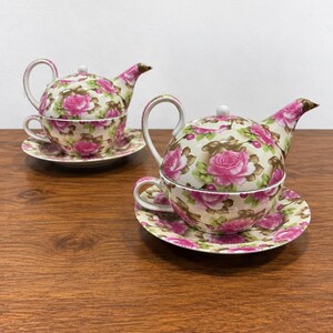 May include: Two tea sets for one, each consisting of a teapot, a teacup, and a saucer. The ceramic pieces feature a cream-colored base decorated with a pattern of vibrant pink roses and green leaves. Each teapot sits securely atop its matching teacup.