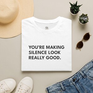 May include: A white short-sleeved t-shirt featuring the black text "YOU'RE MAKING SILENCE LOOK REALLY GOOD." printed on the front. The shirt has a round neckline and a small circular logo on the inside collar area.