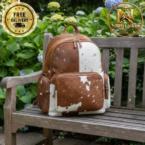 May include: A backpack crafted from cowhide featuring a patchwork pattern of brown and white fur. The bag includes a main zippered compartment, a large front zippered pocket, and two smaller side zippered pockets with brown leather trim.