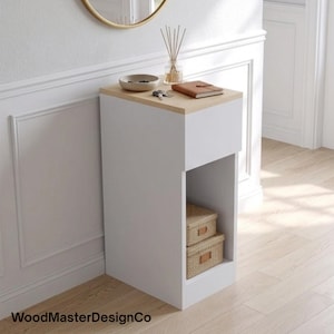 May include: A white rectangular console table with a light wood top surface. The unit features an open lower shelf containing two stacked woven storage boxes. The top surface holds a small bowl, a set of keys, and a brown notebook.