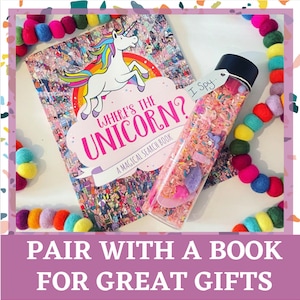 May include: A colorful book titled "Where's the Unicorn? A Magical Search Book" with a unicorn illustration on the cover. A clear plastic bottle filled with colorful sprinkles and small toys is next to the book. The bottle has a black lid with the words "I Spy..." written on it.