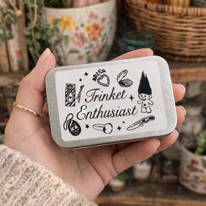 Może przedstawiać: Prostokątne, srebrne pudełko z czarno-białym wzorem graficznym. Projekt zawiera ilustracje butelki perfum, serca, muszli, lalki trolla i innych drobiazgów. Na pudełku wydrukowano napis "Trinket Enthusiast".