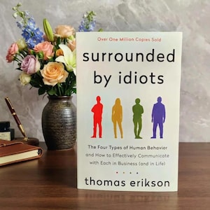 May include: A book titled Surrounded by Idiots by Thomas Erikson with a white cover featuring four colored silhouettes in red, yellow, green, and blue. The text reads Over One Million Copies Sold and The Four Types of Human Behavior.
