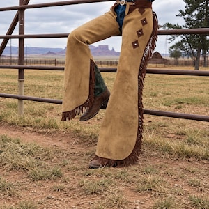 May include: Tan suede leather chaps featuring long brown fringe along the outer legs, a dark brown waistband with a metal buckle, and dark brown diamond-shaped patches with silver studs on the upper thigh area over blue denim jeans.