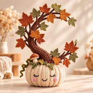 May include: A crocheted cream-coloured pumpkin with a smiling face, closed eyes, and pink cheeks. A brown crocheted tree trunk grows from the top, featuring orange and green maple leaves and a small mushroom at the base of the tree structure.