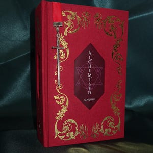Könnte beinhalten: Ein rotes Hardcover-Buch mit goldenen Filigranmustern auf dem Einband. Ein zentrales dunkelrotes Sechseck zeigt den Text ALOHEMISED und SENLINYU mit einem Sternsymbol. Ein rotes Leseband hängt oben herab, befestigt an einem silbernen Schwertanhänger.