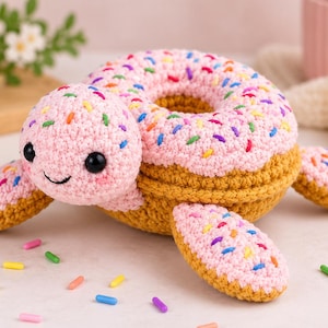 May include: A crocheted turtle plush toy featuring a shell shaped like a pink frosted donut with colorful sprinkles. The turtle has a light pink head with black safety eyes, a stitched smile, and golden-brown yarn for the body and flippers.