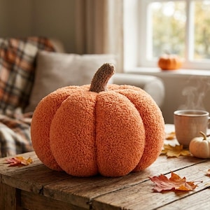Puede incluir: Un cojín grande y redondo con forma de calabaza, con una superficie texturizada y difusa, y un tallo marrón de punto. El cojín está colocado sobre una superficie de madera junto a una taza de cerámica y dos calabazas naturales pequeñas.