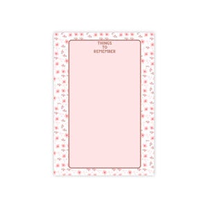 Peut inclure: Un bloc-notes rectangulaire avec un centre rose clair et une bordure ornée d'un motif de petites fleurs roses et de feuilles vertes. Le texte "THINGS TO REMEMBER" est imprimé en marron en haut au centre de la zone rose, encadrée par un trait marron.